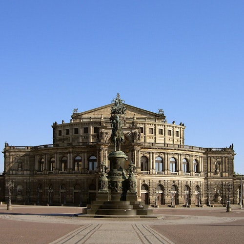 Historic building with ornate architecture, large arched windows, and a central statue set in a spacious plaza under a clear blue sky.