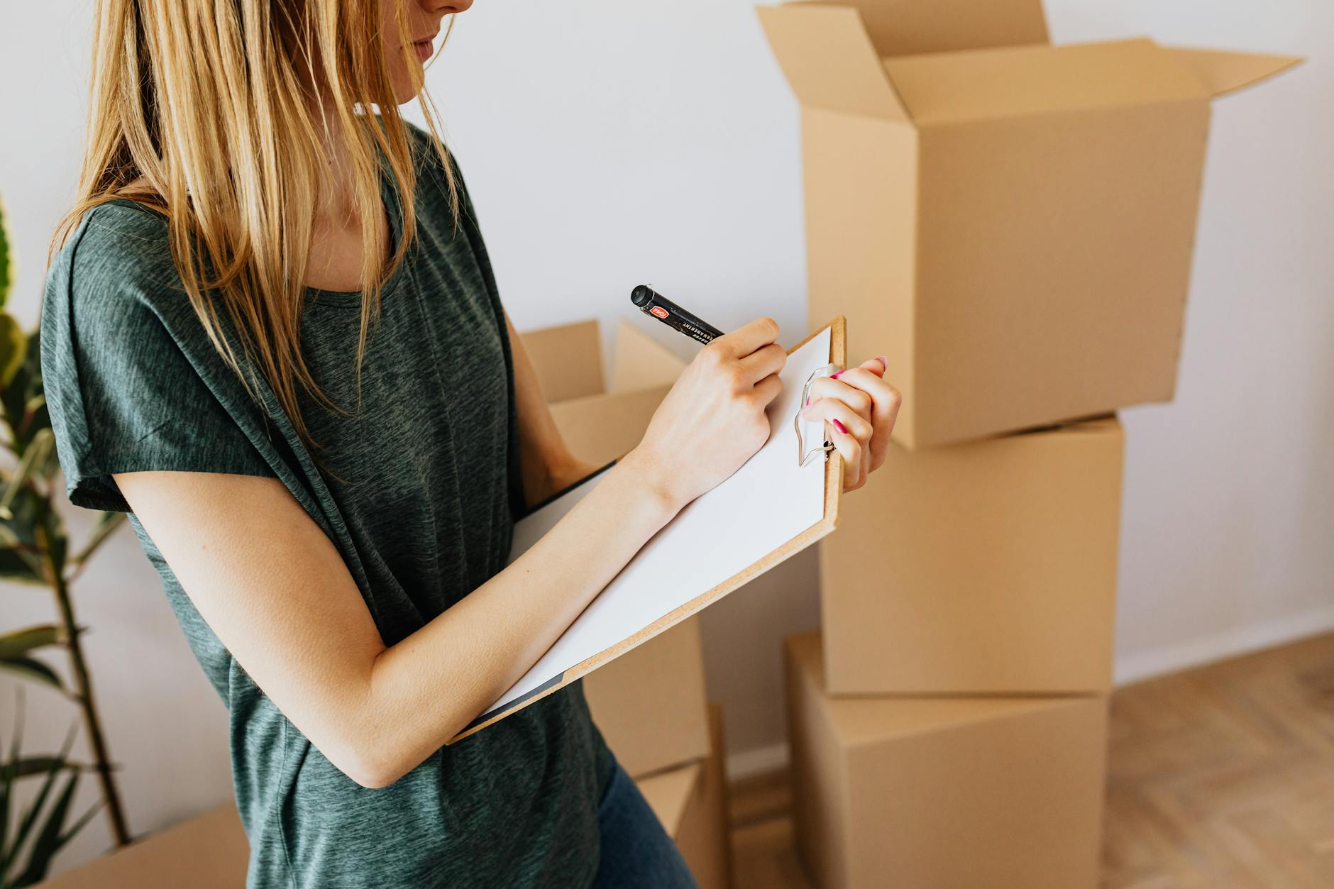 Blonde woman in a green T-shirt writing on a clipboard in front of stacked cardboard moving boxes