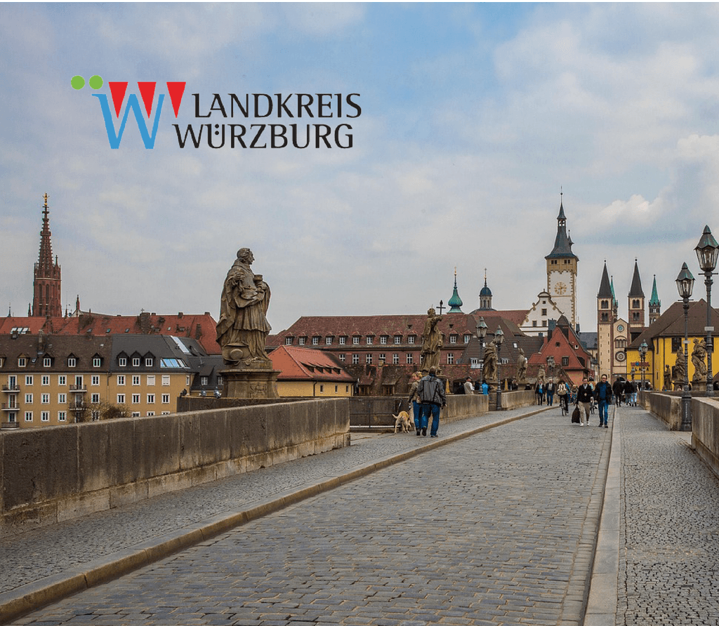 Eine alte Brücke in Würzburg mit vielen älteren typischen Gebäuden für eine Altstadt und einer Kirche, darauf befindet sich das Logo des Landkreis Würzburg
