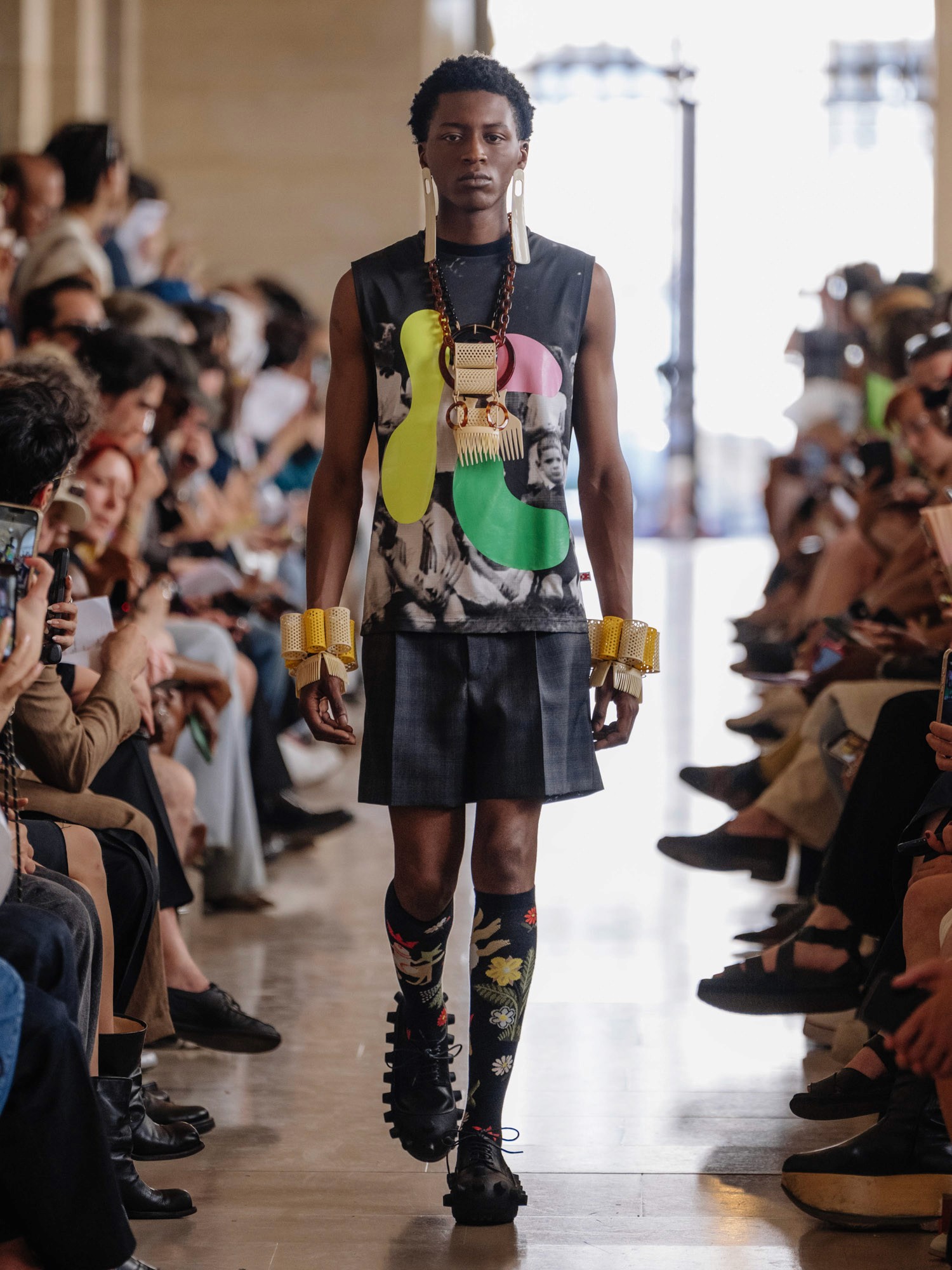 A model walks down the runway in a sleeveless, abstract-patterned top and a dark skirt, accessorized with oversized earrings, bold necklaces, large bangles, and decorative socks paired with unique black shoes, in a fashion show setting filled with seated spectators.