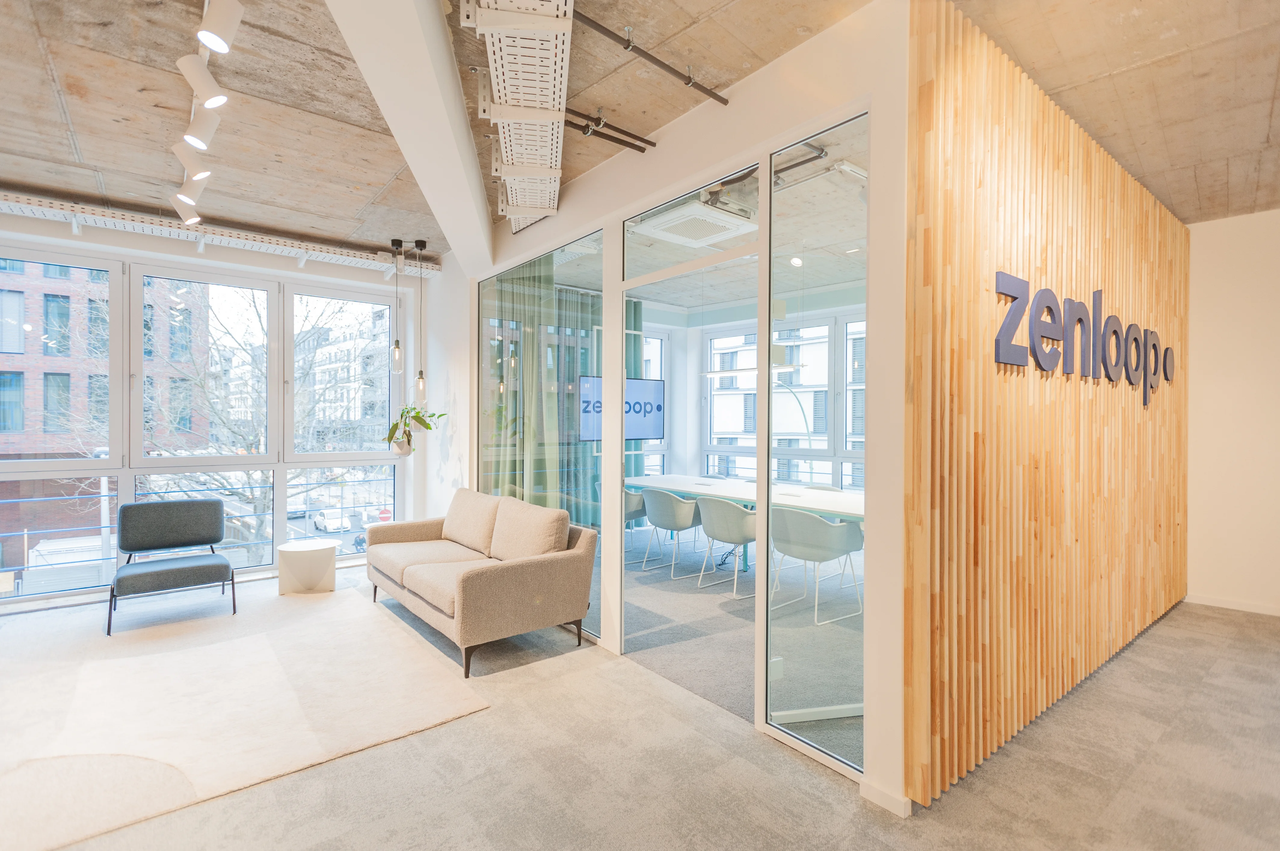 Lounge zone at ZENLOOP with a beige sofa and dark grey armchair by the windows, next to a glazed meeting box clad in vertical wooden slats with the navy zenloop logo.