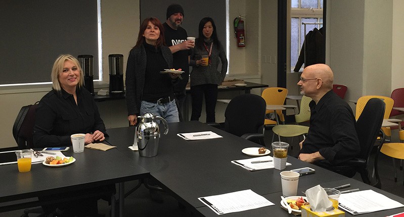 A photo of a group of people sitting in an office room at a table. On it there are some plates with food, some beverages and notepads with pens.