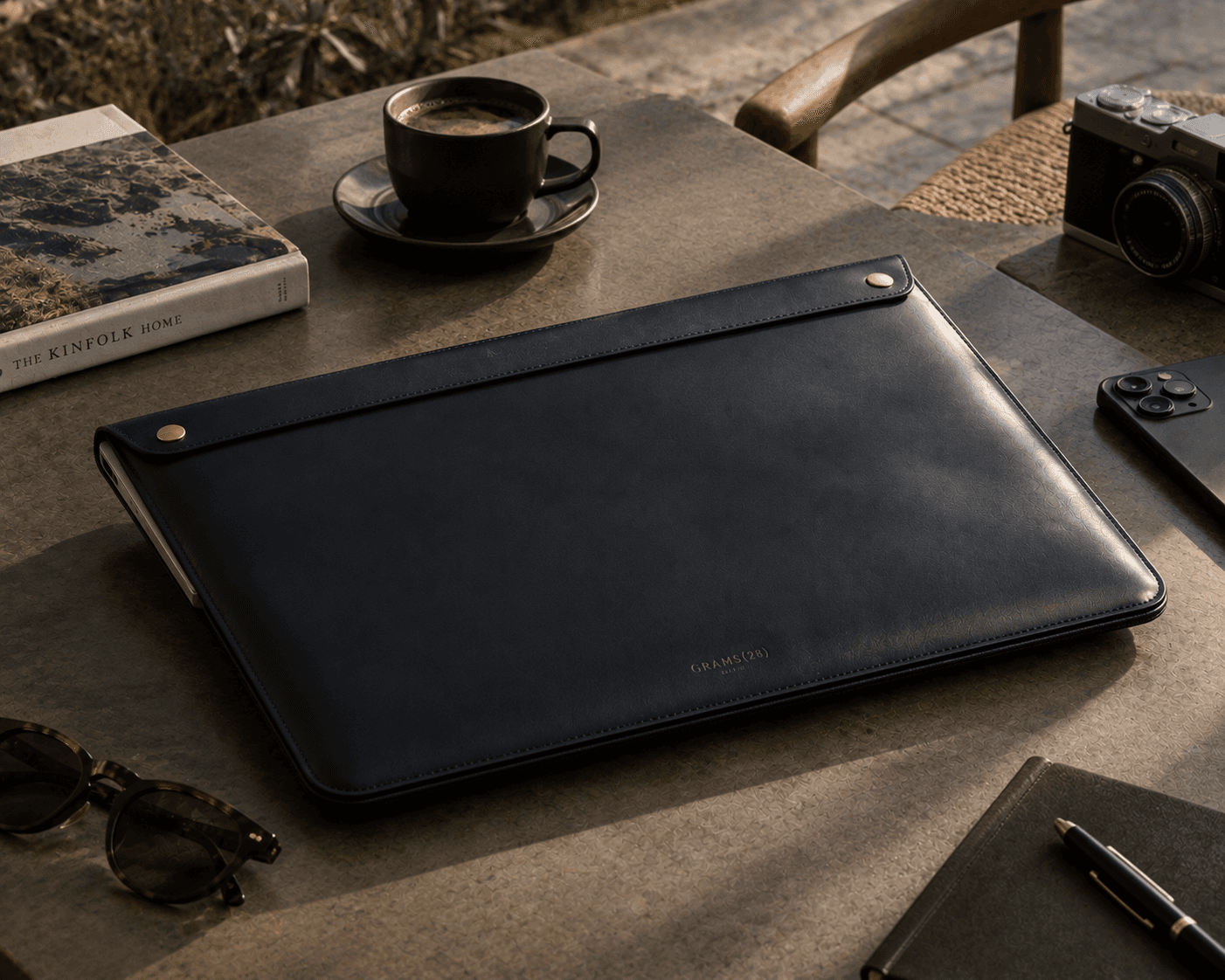A sophisticated tech lifestyle flatlay on a gray concrete outdoor patio table in warm late-afternoon sunlight. At the center remains a black leather laptop folio case with a top flap secured by two brass snap closures and subtle GRAMS(28) branding. Surrounding accessories include a dark espresso cup on a saucer, a vintage rangefinder camera, a dark smartphone, an olive green leather notebook with a metallic pen, tortoiseshell sunglasses, and a copy of "THE KINFOLK HOME" book alongside a woven wooden chair in the background.