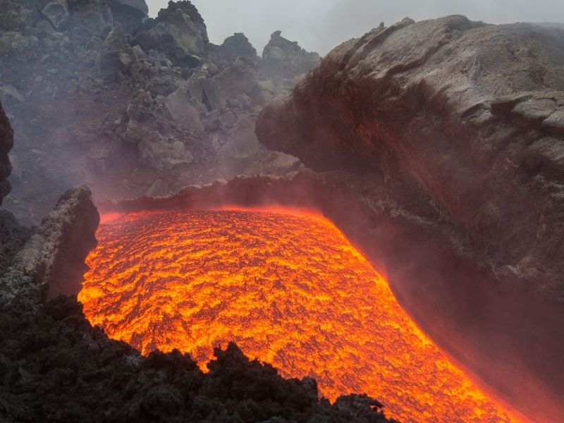 Glowing lava flowing steadily from Mount Etna during an effusive eruption, with molten rock moving slowly between dark volcanic formations.