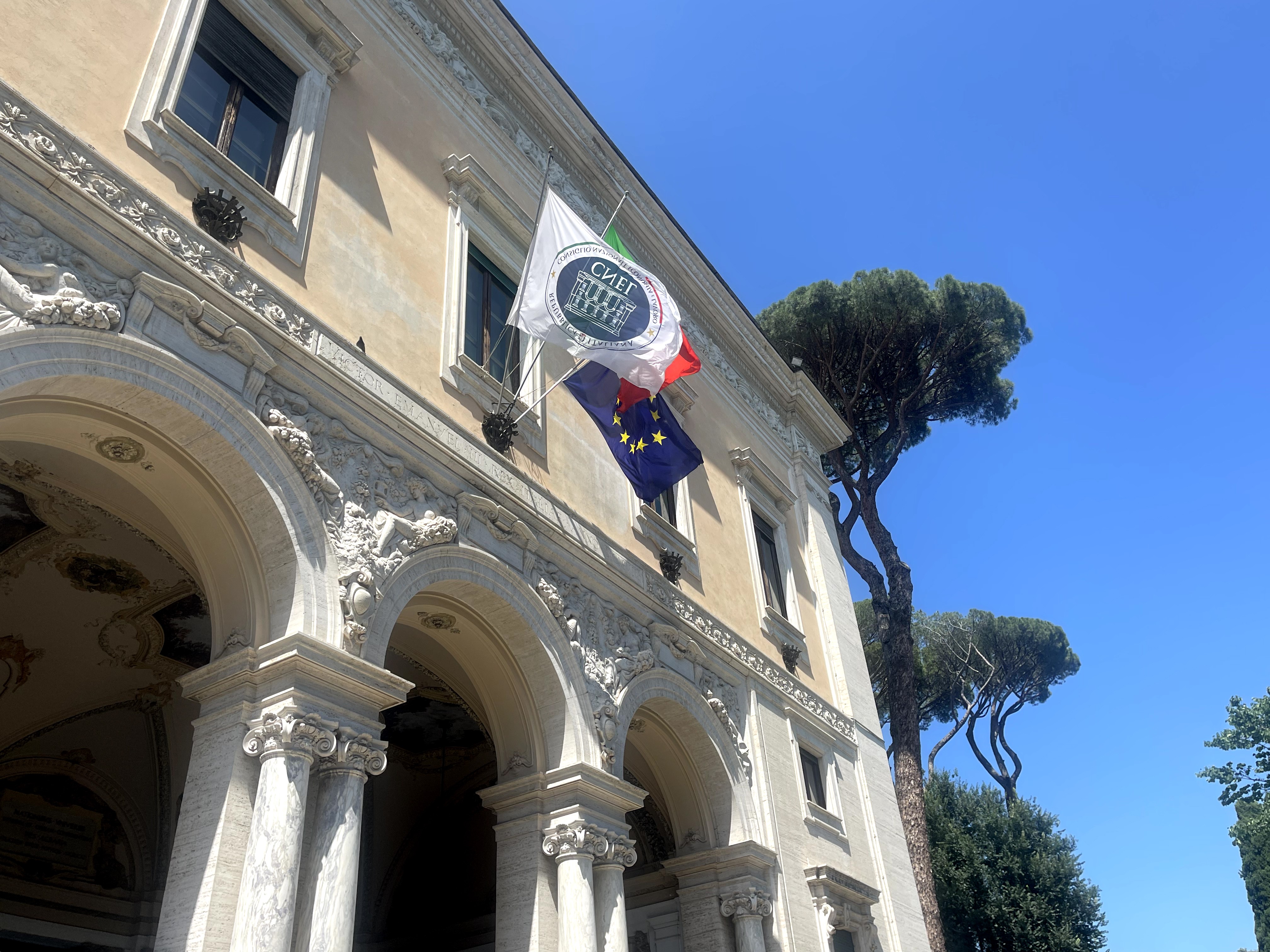 Facciata monumentale di Villa Lubin a Roma, sede del CNEL, con bandiere italiana ed europea su sfondo di cielo sereno e alberi secolari.
