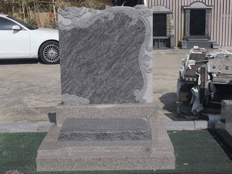 A grey granite tombstone with cloud and wave carvings on the top left corner. The tombstone is placed outdoors, reflecting traditional design elements.