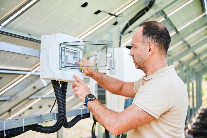 Eine Person in einer Sicherheitsweste hält ein Smartphone in der Nähe eines Solarpanels, mit klarem Himmel im Hintergrund.
