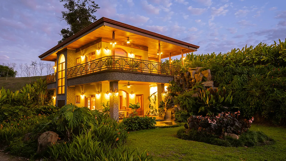 A two-story Arenal Hills villa illuminated at dusk, with a covered balcony and lush tropical gardens.