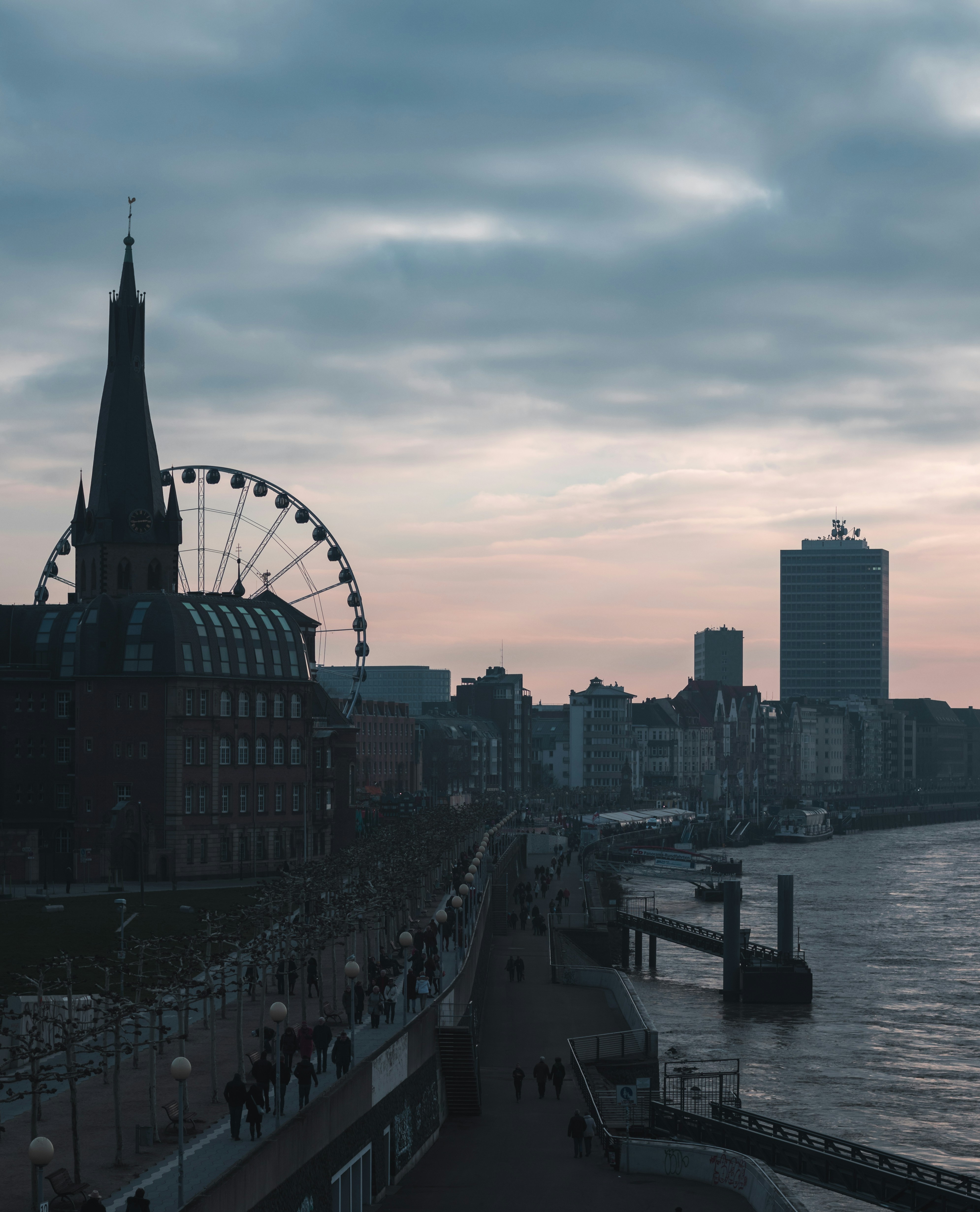 Das Bild zeigt die Düsseldorfer Rheinuferpromenade mit der St. Lambertus Basilika und einem Riesenrad im Hintergrund.