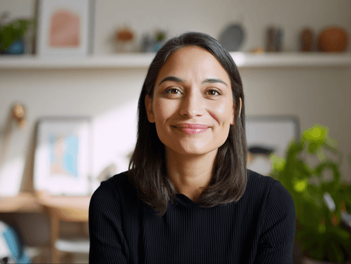 A woman with long dark hair smiles warmly at the camera. Behind her, blurred shelves hold decor and plants, suggesting a cozy home setting.