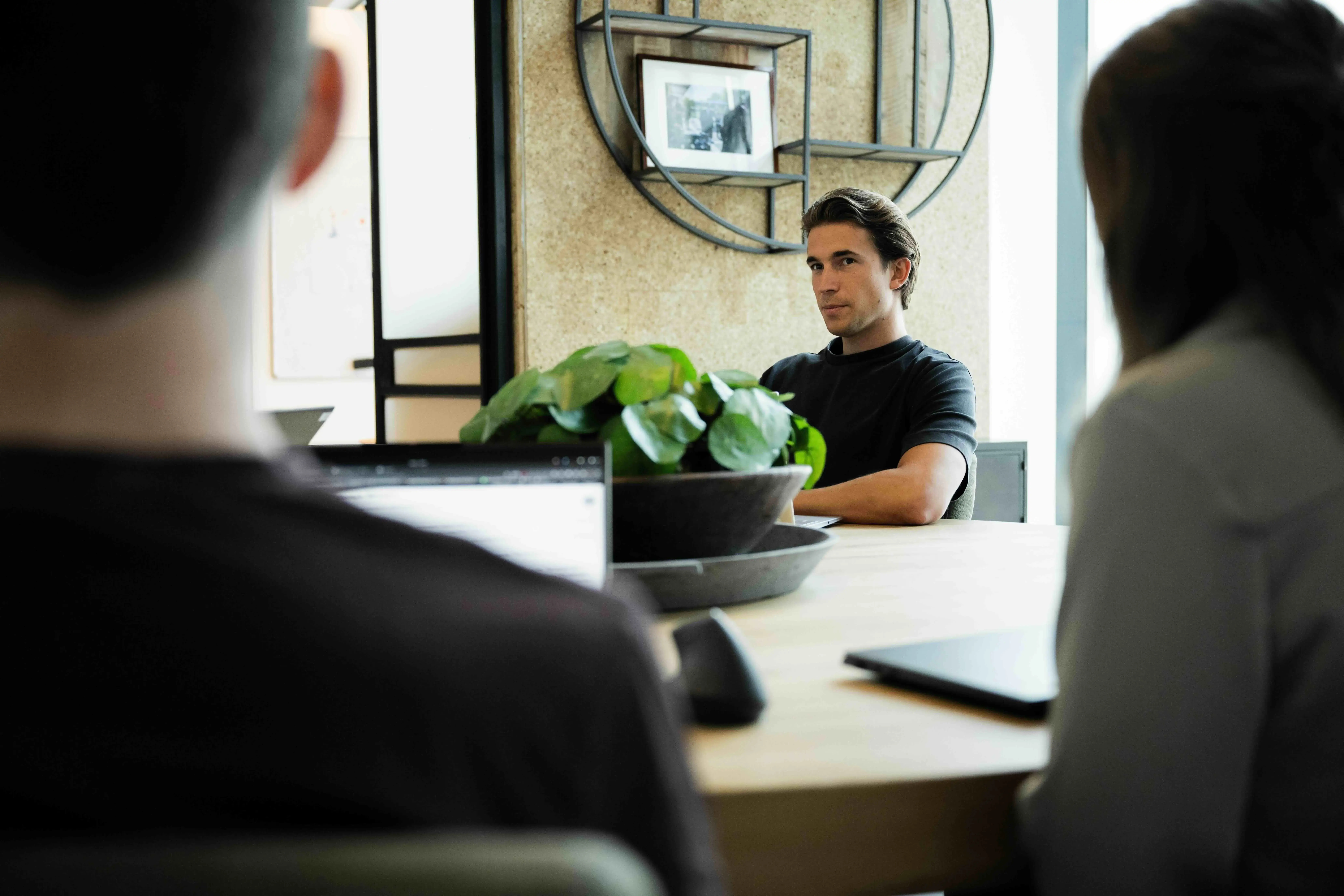 In a modern office setting, a group of individuals is engaged in a meeting around a large wooden table, with a laptop in the foreground and a central decorative plant enhancing the contemporary workspace ambiance.