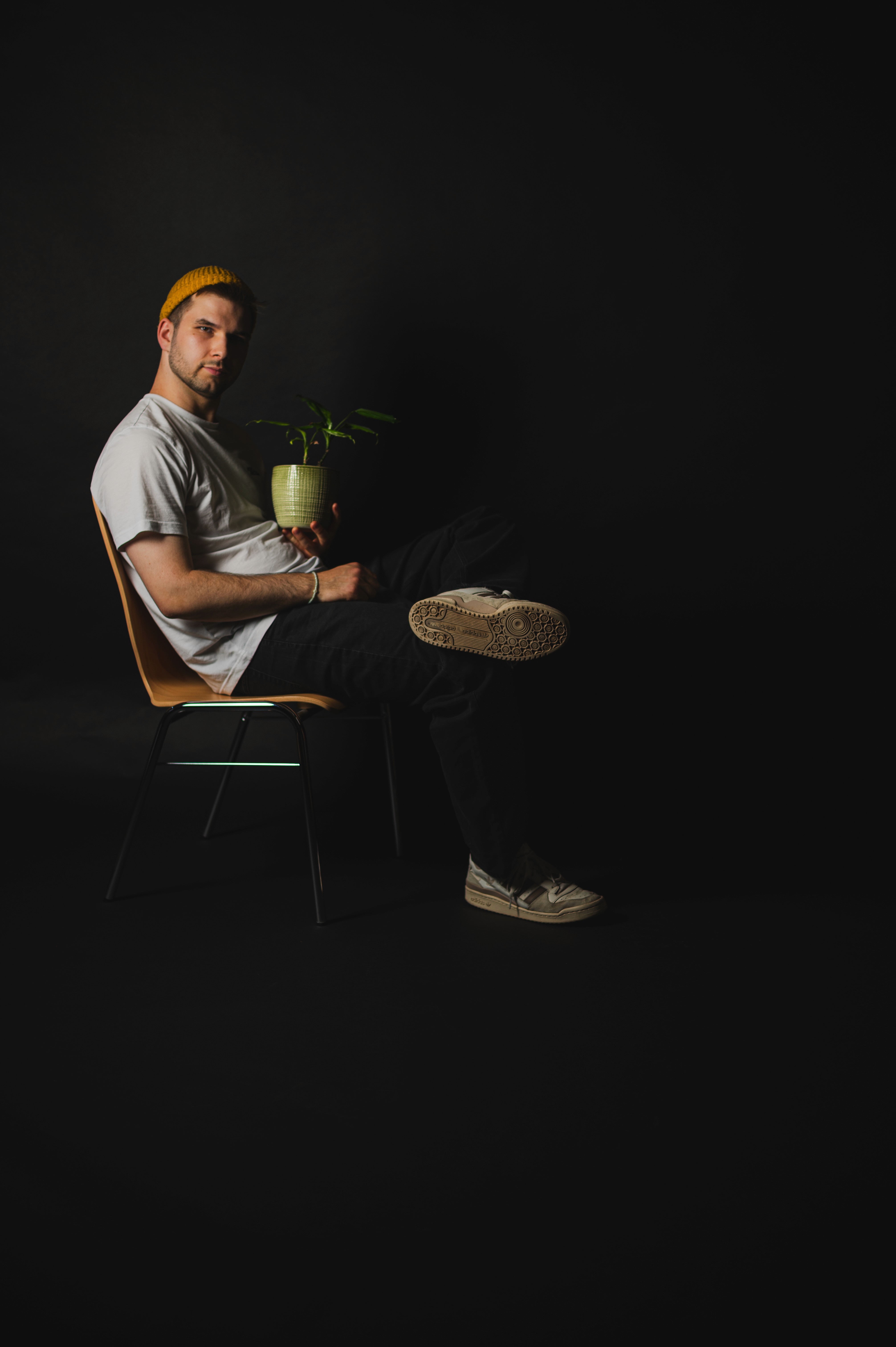 Ein Mann sitzt auf einem Stuhl vor schwarzem Hintergrund, trägt eine Kappe und hält eine Topfpflanze auf dem Schoß.