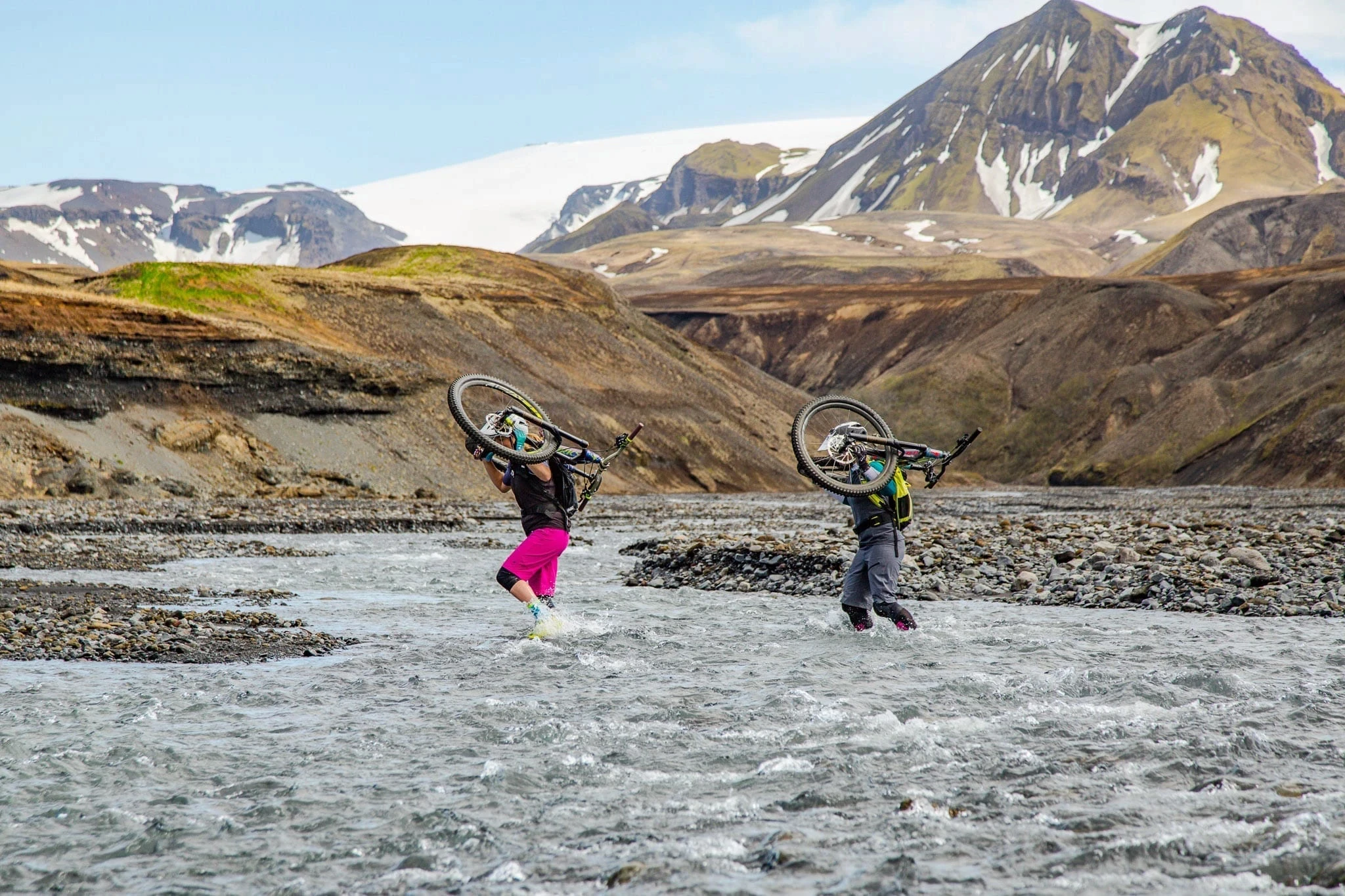 Two cyclists carrying bikes through a glacial river with mountains behind.