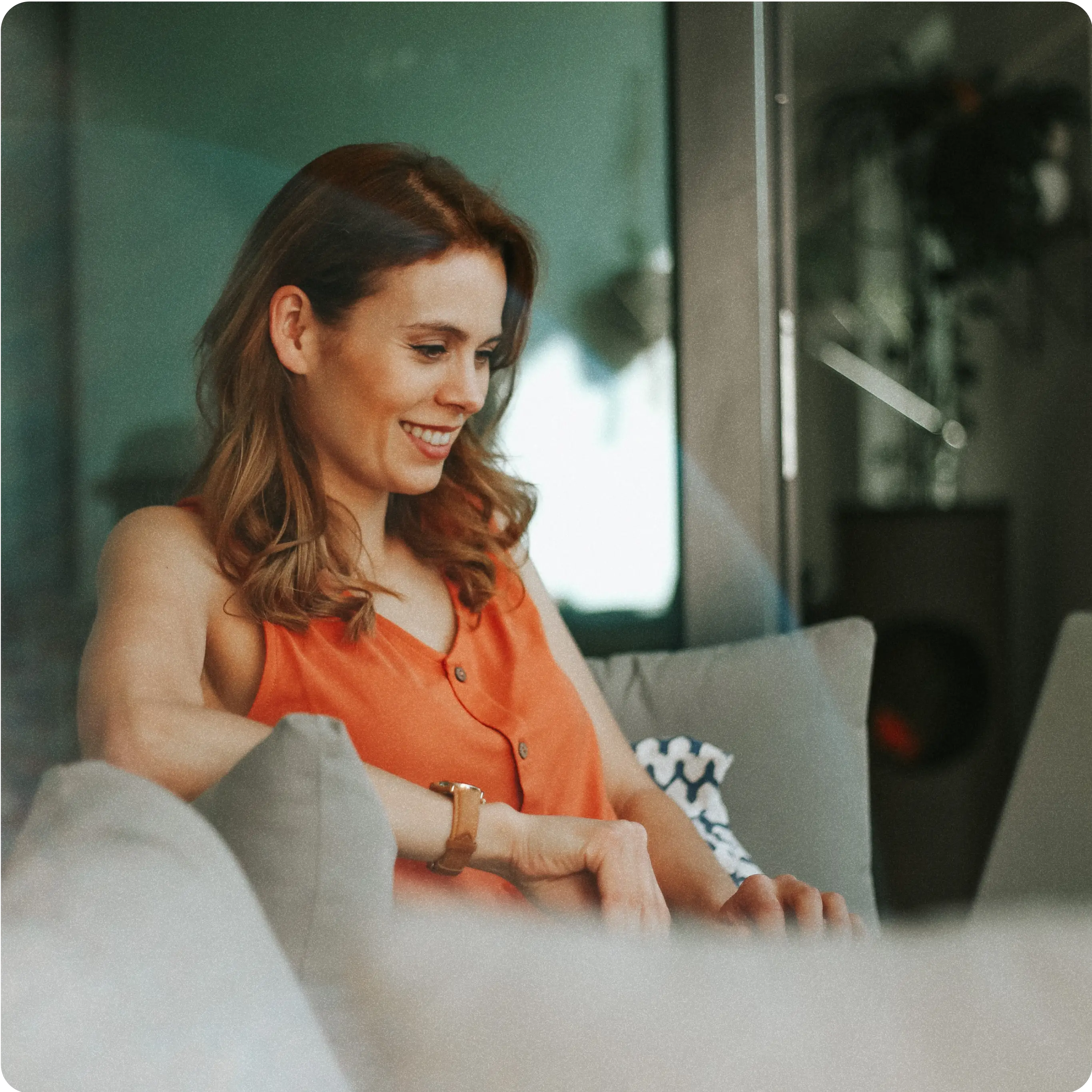 A woman with auburn hair, wearing an orange sleeveless top, is sitting on a comfortable couch. She is smiling and looking at something in front of her, possibly a laptop or tablet, suggesting she is engaged in an activity or enjoying a moment of relaxation. The background shows a modern, cozy living space with soft lighting and a few decorative elements.