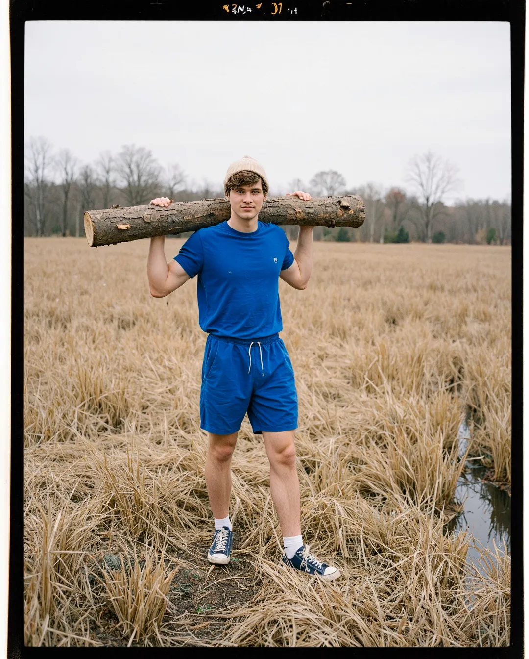 Person in bright blue athleisure carrying wooden log across golden autumn wetland with bare trees in background, film-style photograph
