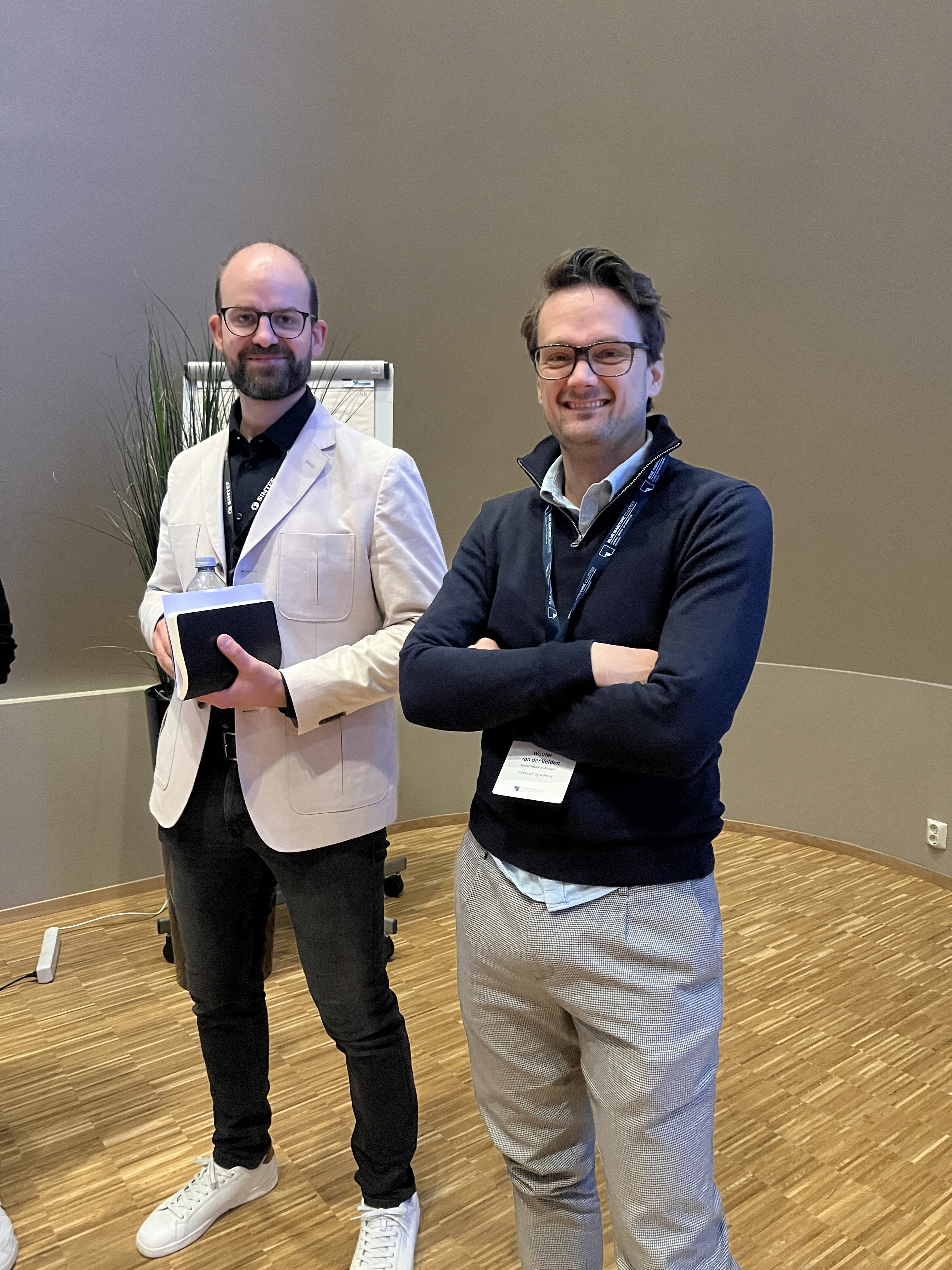 Two men stand together, one holding a tablet and the other crossing his arms, against a neutral background.