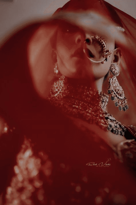 Moody close-up of an Indian bride in elaborate jewelry, with soft red hues creating a dramatic, mysterious effect. Perfect example of moody and dramatic wedding photography capturing the bride's elegance and intensity.