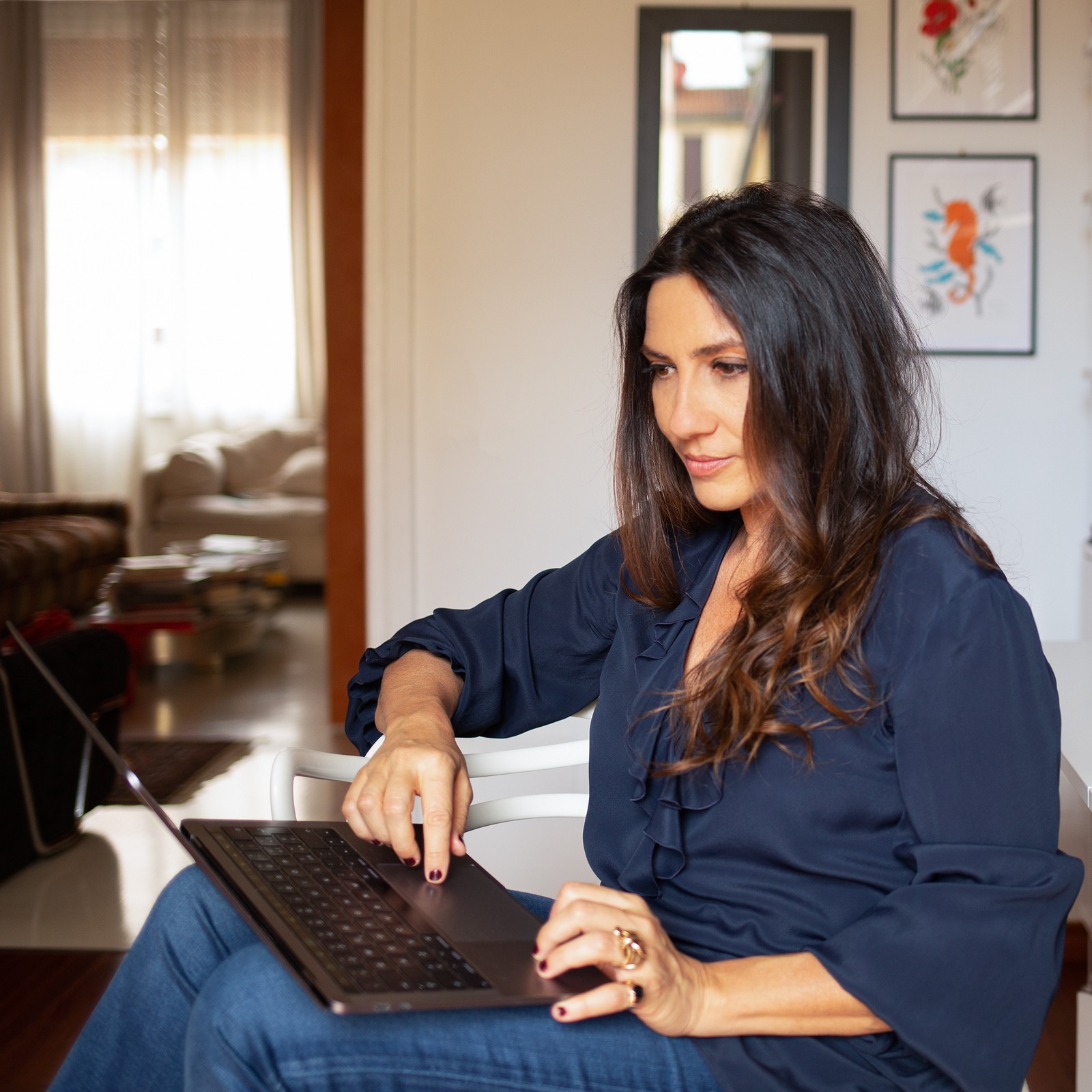 A young person with curly hair smiles at the camera while sitting in front of a laptop against a dark background.
