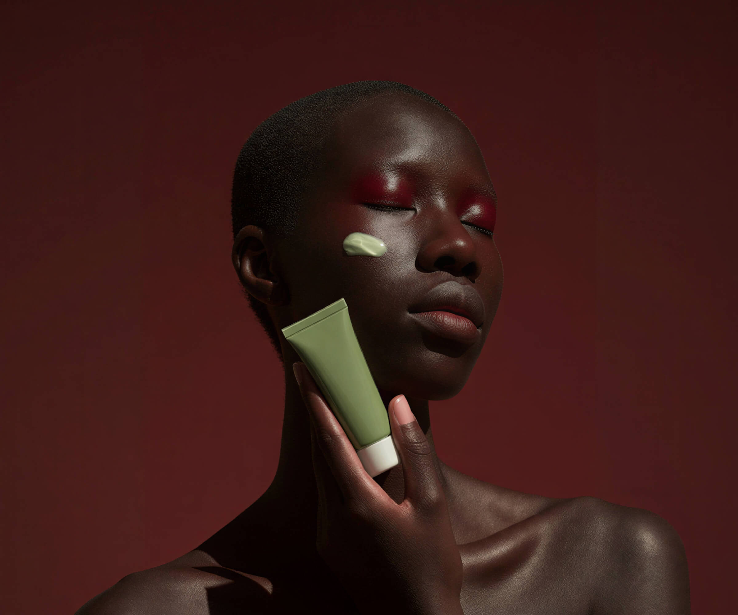 A person with dark skin is gently holding a green object near their face against a deep red backdrop.