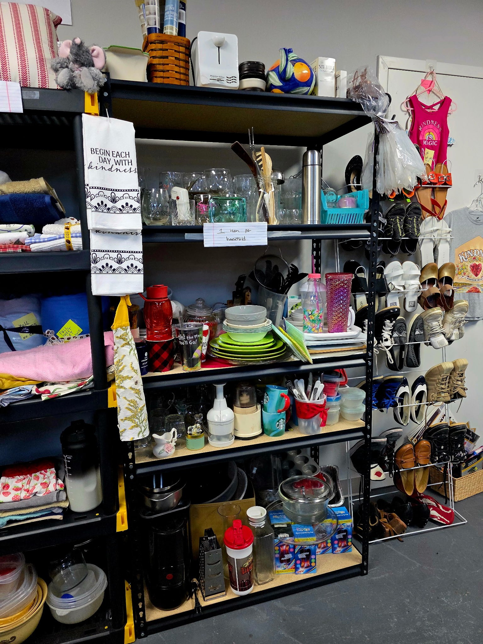 Shelves stacked with donated kitchenware, dishes, small appliances, and household items next to a shoe rack inside the Clothed in Kindness store in Jefferson County, Indiana.