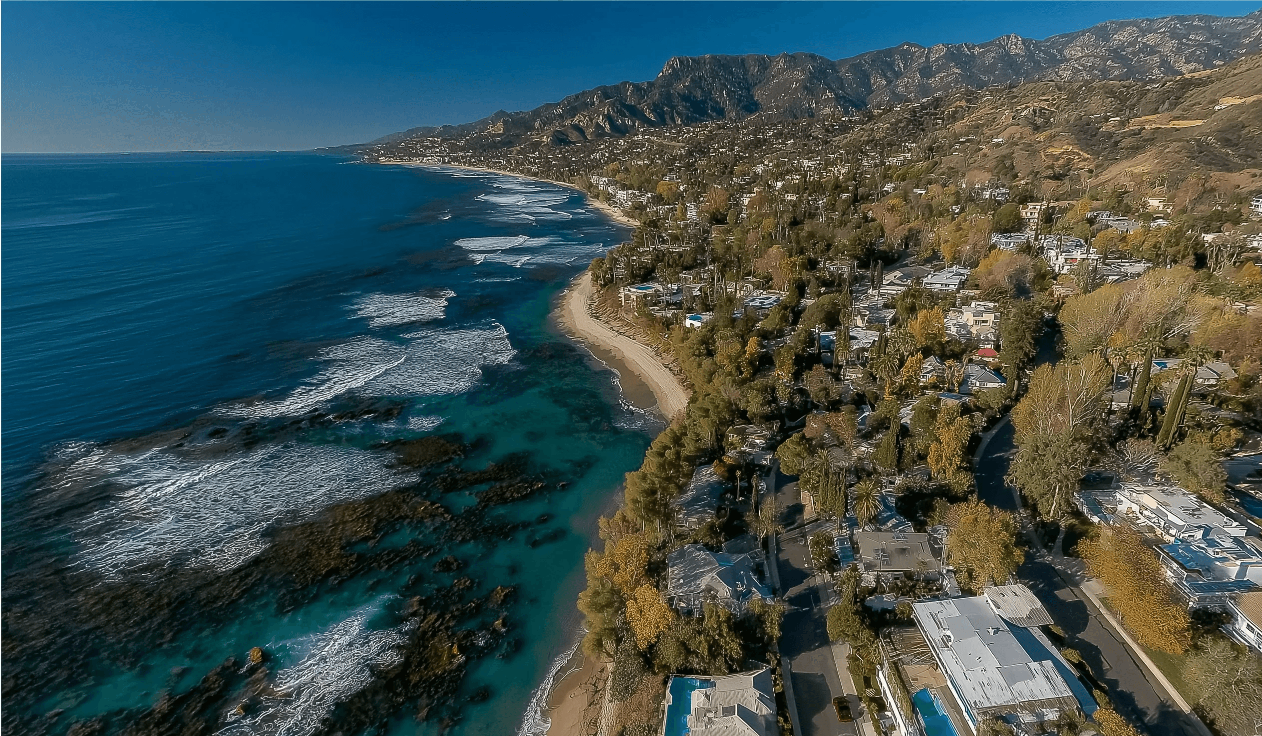 High-angle drone shot of a beautiful coastline featuring ocean waves crashing onto a sandy beach, bordered by a hillside community of luxury homes.