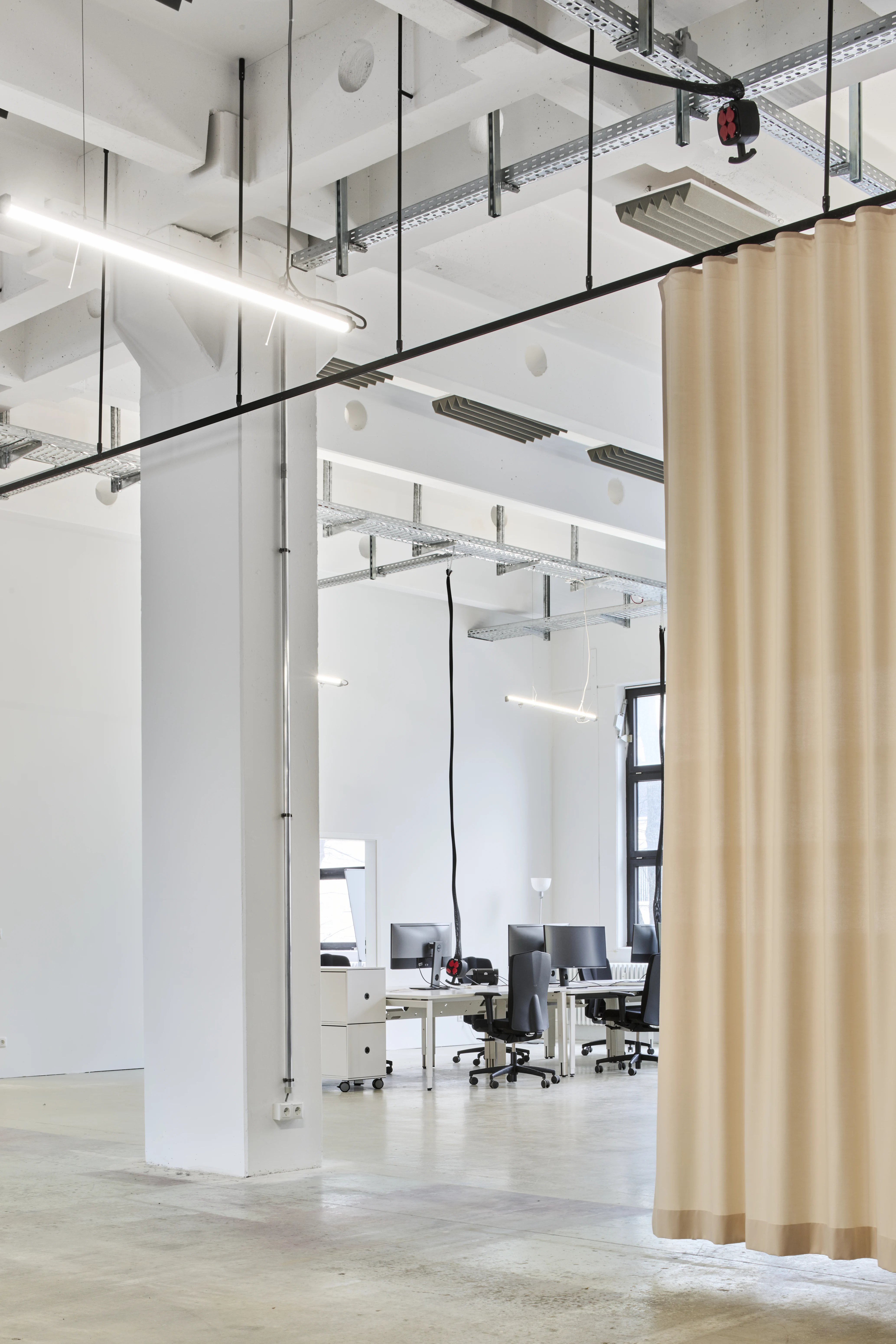 View into the office zone of the FLACONI CONTENT STUDIO with white desks and black monitors behind a beige ceiling-mounted curtain in an industrial white loft.