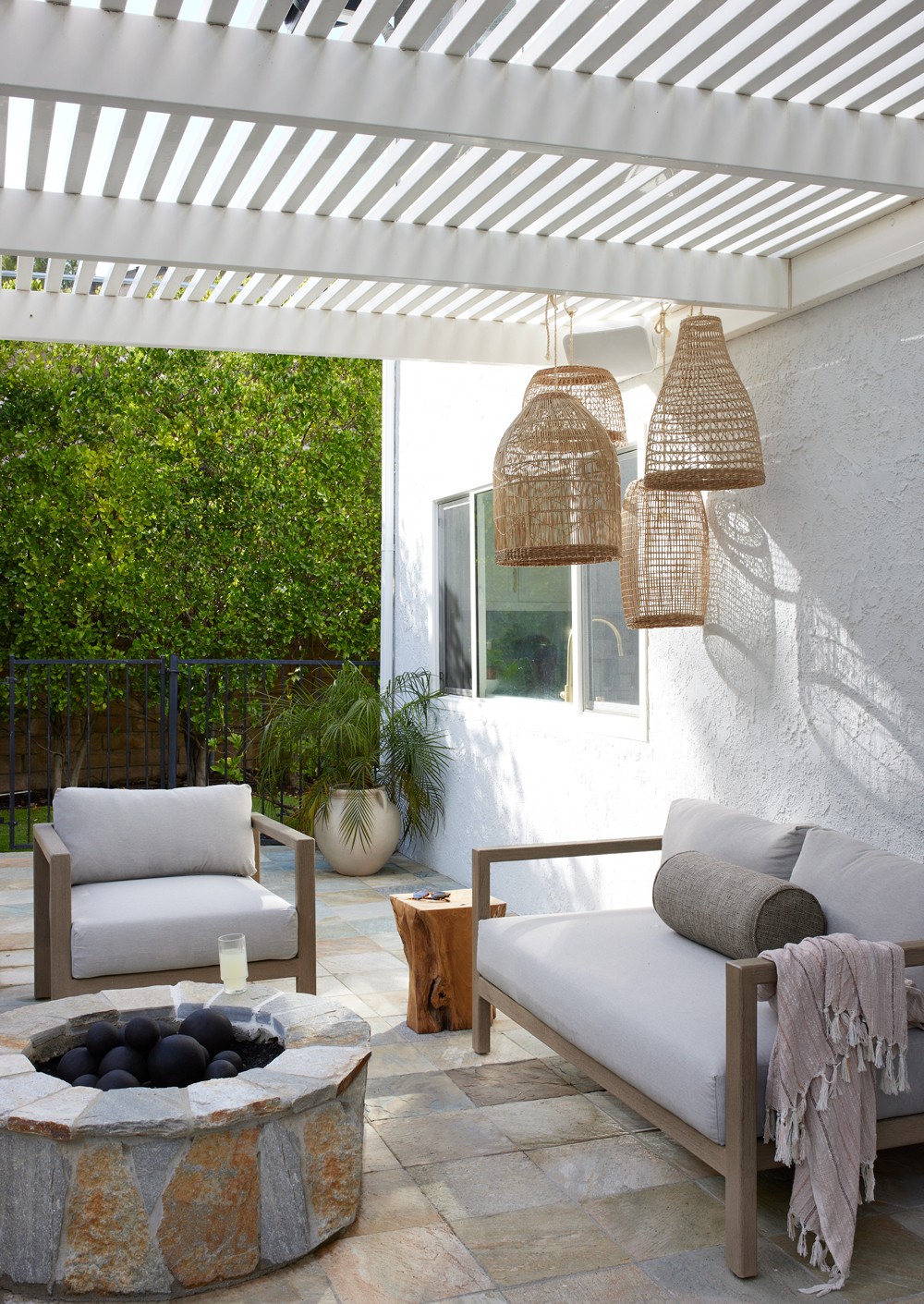 This tranquil gathering spot features earthy tones, a wooden overhead structure, and a stone fire-pit, complemented by wicker accents and sectional seating for a cozy, shaded escape