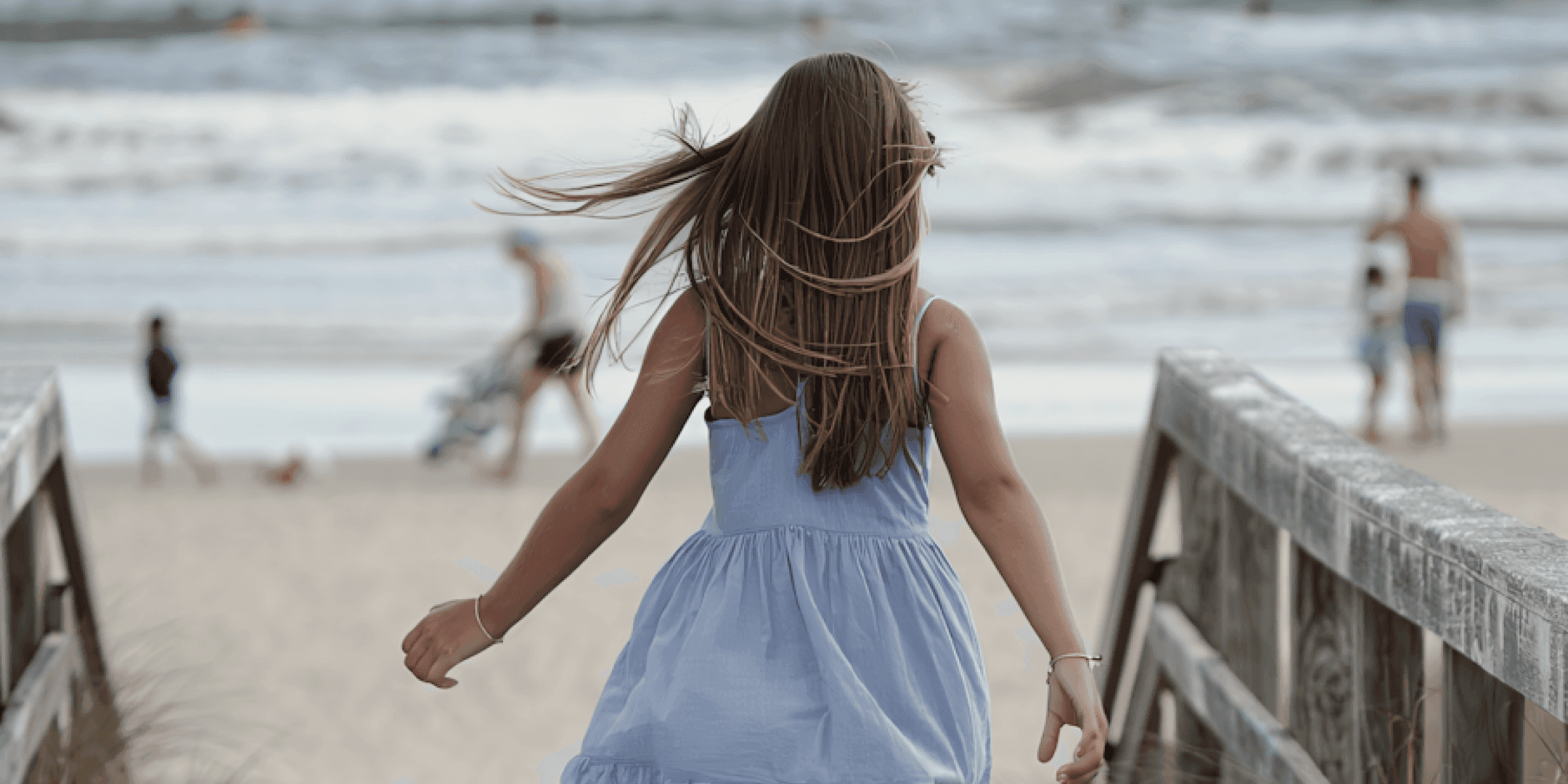 Girl in blue dress walking toward the beach with waves and people in background