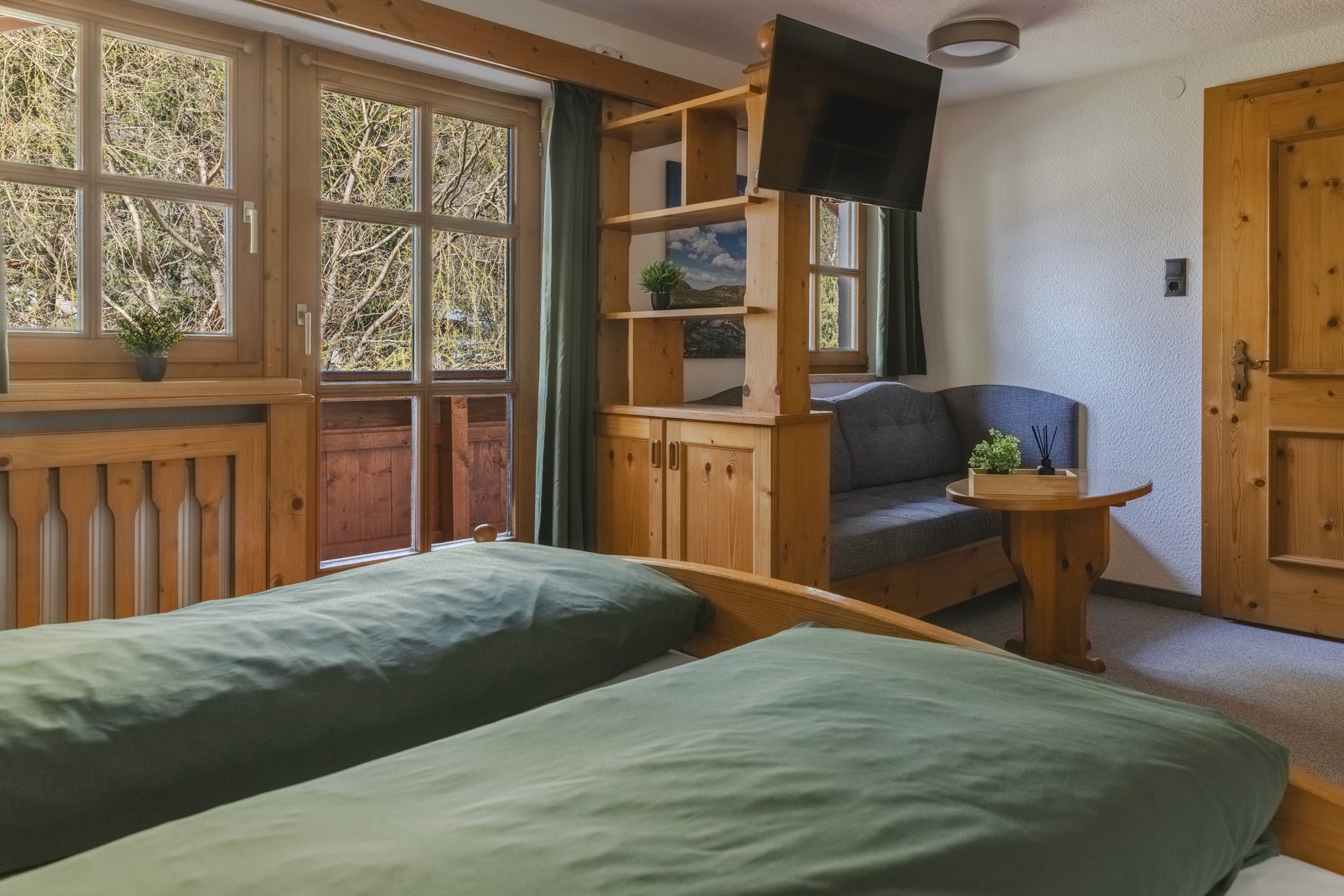 Ein gemütliches, rustikales Schlafzimmer verfügt über ein Doppelbett aus Holz mit grünen Tagesdecken, eine offene Balkontür mit sonnigem Blick nach draußen, einen an der Wand montierten Fernseher und kleine Topfpflanzen für einen Hauch von Grün.
