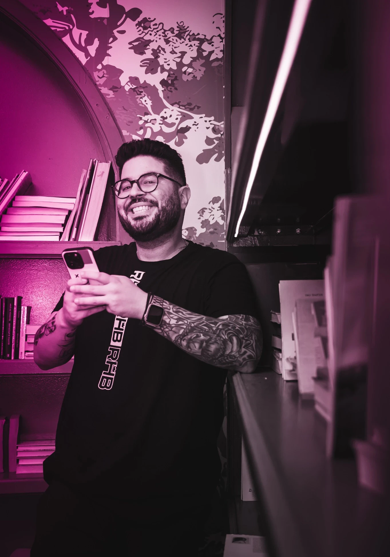 John Demelis, tattooed arms smiles while holding a phone. He is wearing a black "RHB DRIP" t-shirt and leaning against a bookshelf.