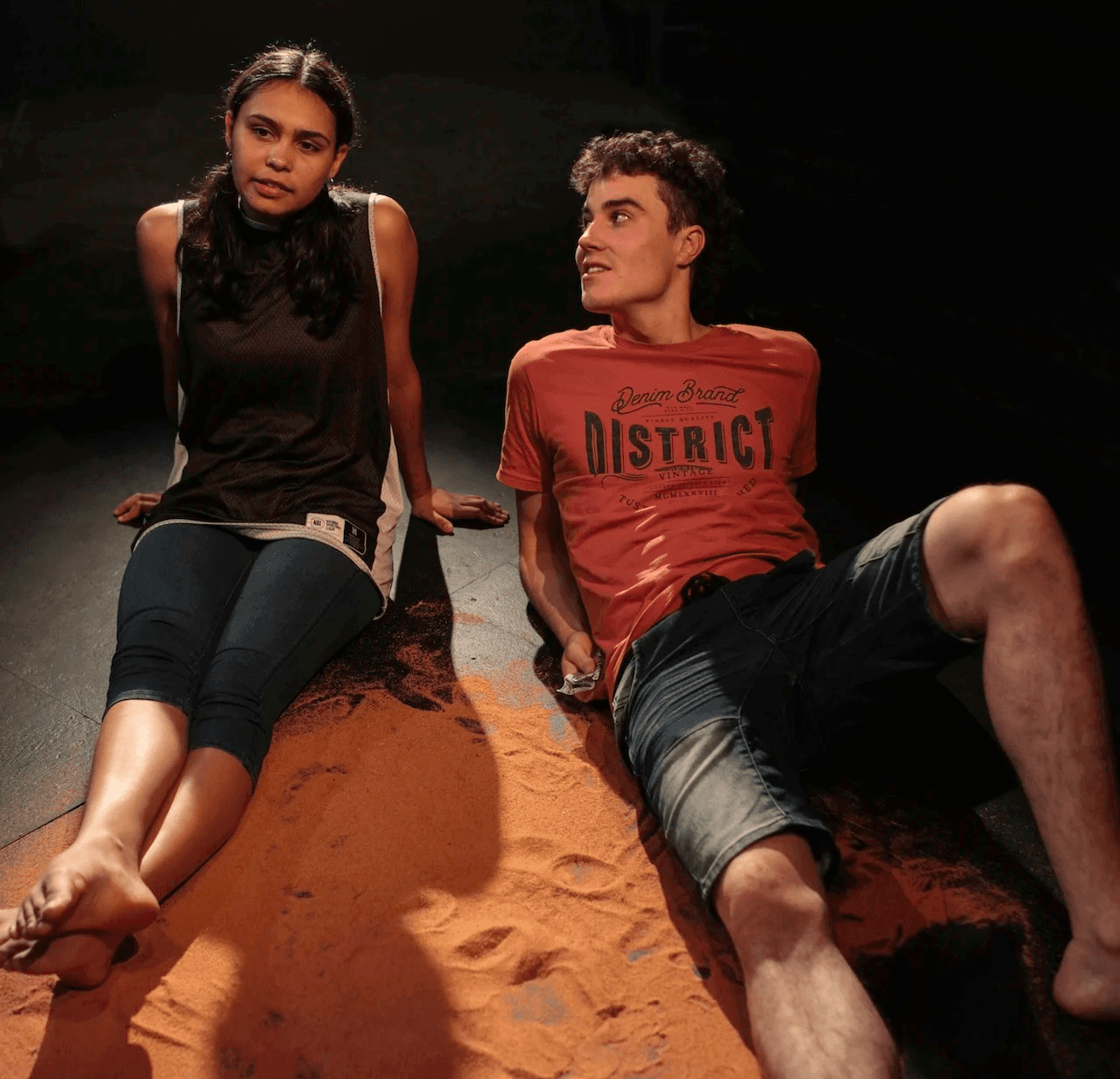 Two young people sit barefoot on a stage covered with red earth. The girl looks thoughtful with her hands behind her, while the boy reclines next to her, looking up at her. They appear deep in conversation under low, warm lighting.
