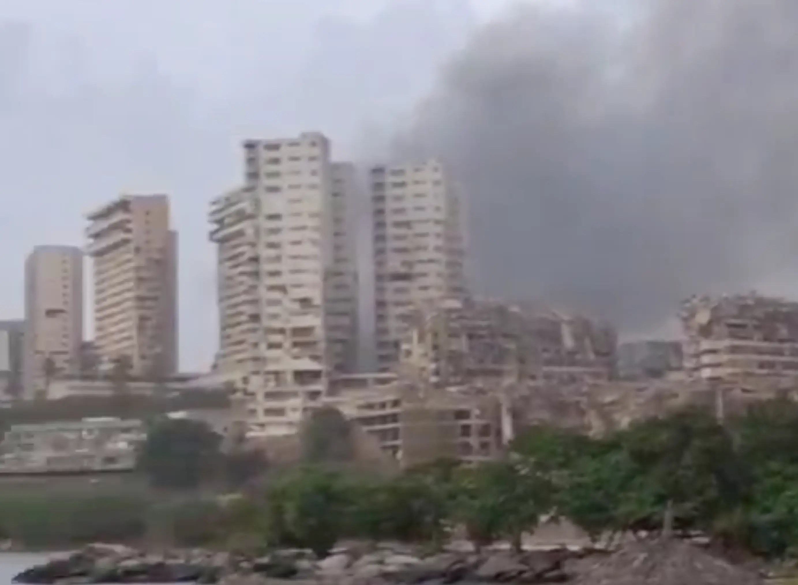 Smoke rises behind damaged high-rise buildings in an urban area, with trees and rubble in the foreground.