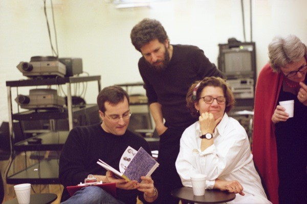 A photo of three people checking a sketchbook while in the background some projectors and tv sets are stacked on tables.
