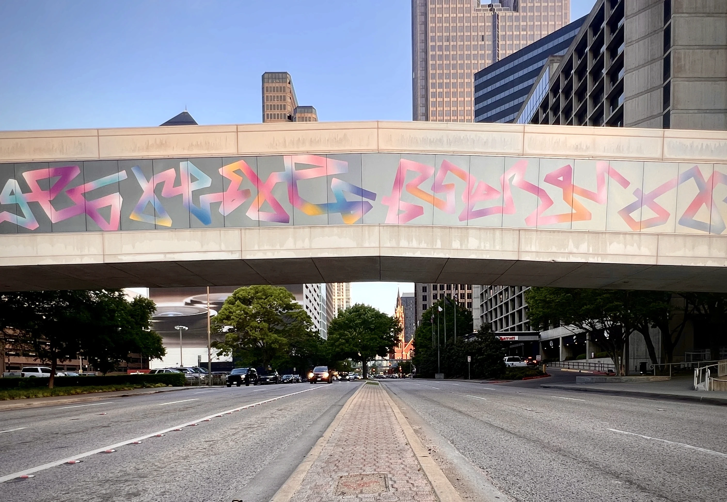 A wide shot of a street under a colorful mural on a bridge, with buildings visible in the background.