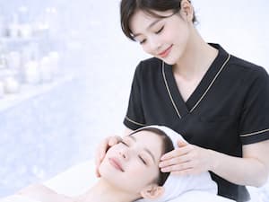 目を閉じてリラックスする女性に、優しくフェイシャルマッサージを施すエステティシャン