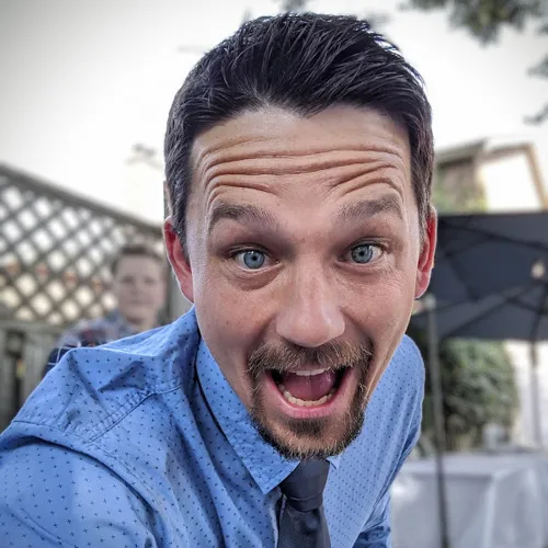 A cheerful person smiling at the camera during an outdoor event, dressed in a light blue shirt and tie, with a lattice backdrop and greenery in the background