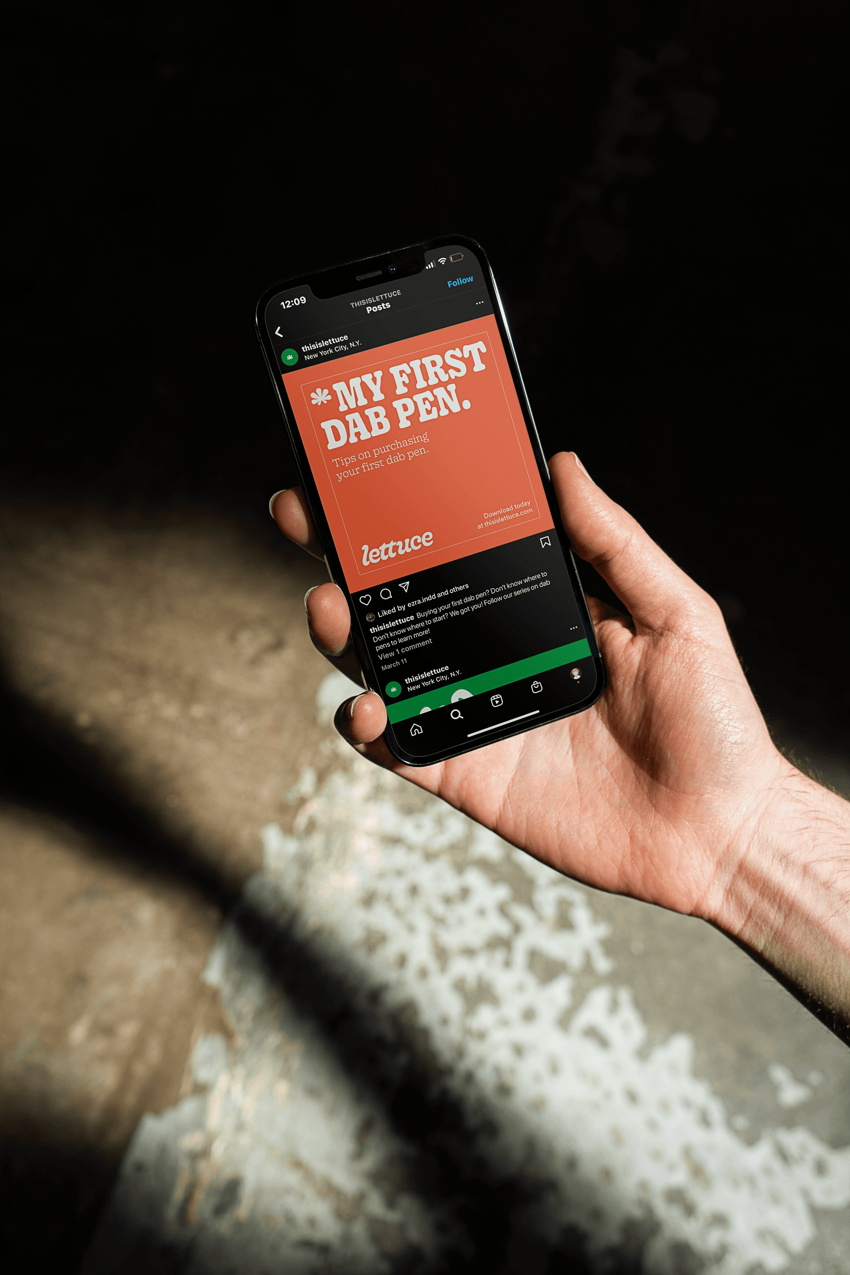 A hand holds a smartphone displaying the LETTUCE app. The screen shows an orange banner with the text “MY FIRST DAB PEN.” and smaller text below. The app interface includes icons for home, settings, news, contacts, messages, and profile. The phone is held against a dark background with a shadow cast on the lower left. The background also shows a textured surface with light and dark areas.