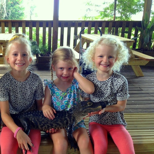 三個年輕女孩，兩個穿著豹紋上衣和粉紅褲子，坐在有野餐桌的木甲板上，手裡抱著一隻小鱷魚。