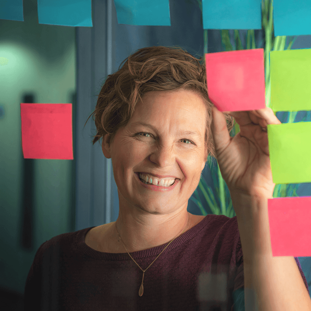 Frau mit kurzem gewelltem Haar und Pullover steht vor einer Glaswand mit mehreren bunten Postits