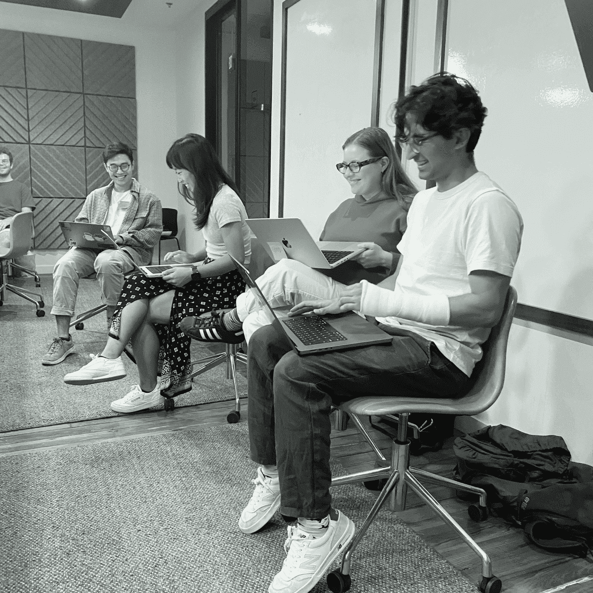 Four young adults, two men and two women, sit in chairs in a modern office setting, each looking at their laptops, with one man in the foreground wearing a cast on his arm.