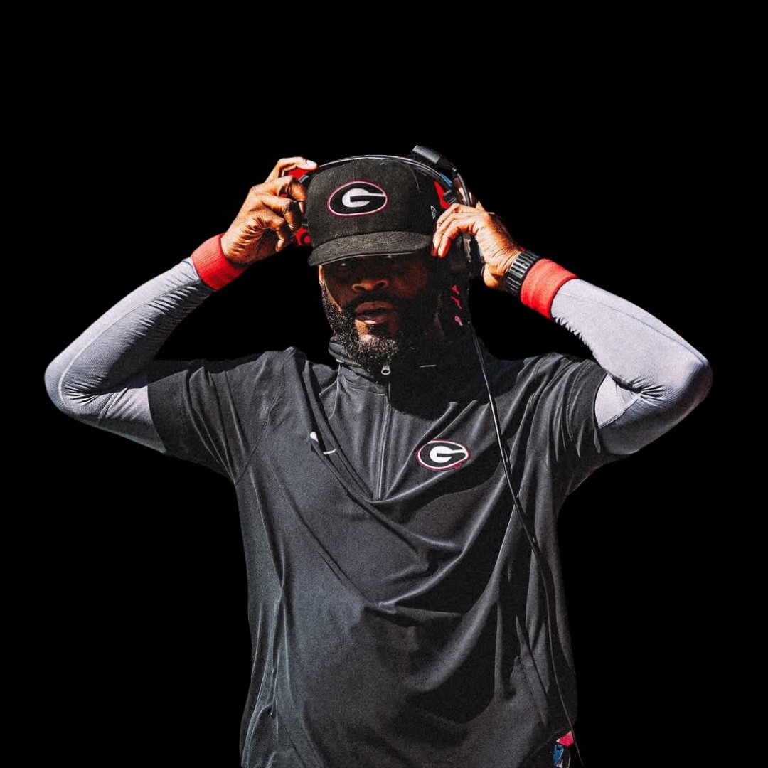 Larry Knight wearing a black Georgia Bulldogs cap and headset adjusts his headphones. He is dressed in a black athletic shirt with the teams logo and stands against a black background.