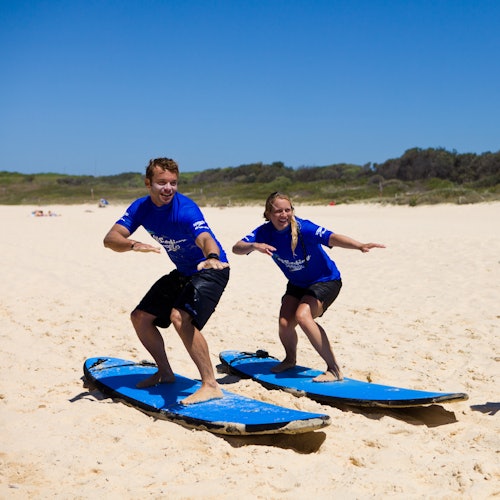 Dalawang tao na naka-asul na kamiseta ang nagsasanay ng mga pusisyon sa pagsu-surf sa asul na surfboard sa mabuhanging dalampasigan na may luntiang tanawin sa likuran.