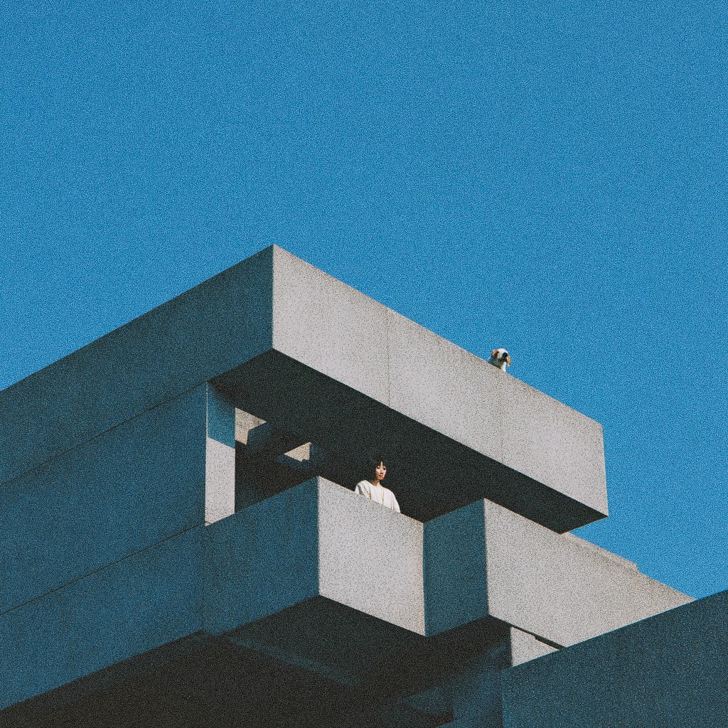 A person stands on a balcony of a brutalist concrete building, with another figure on the top ledge, both framed against a bright blue sky.