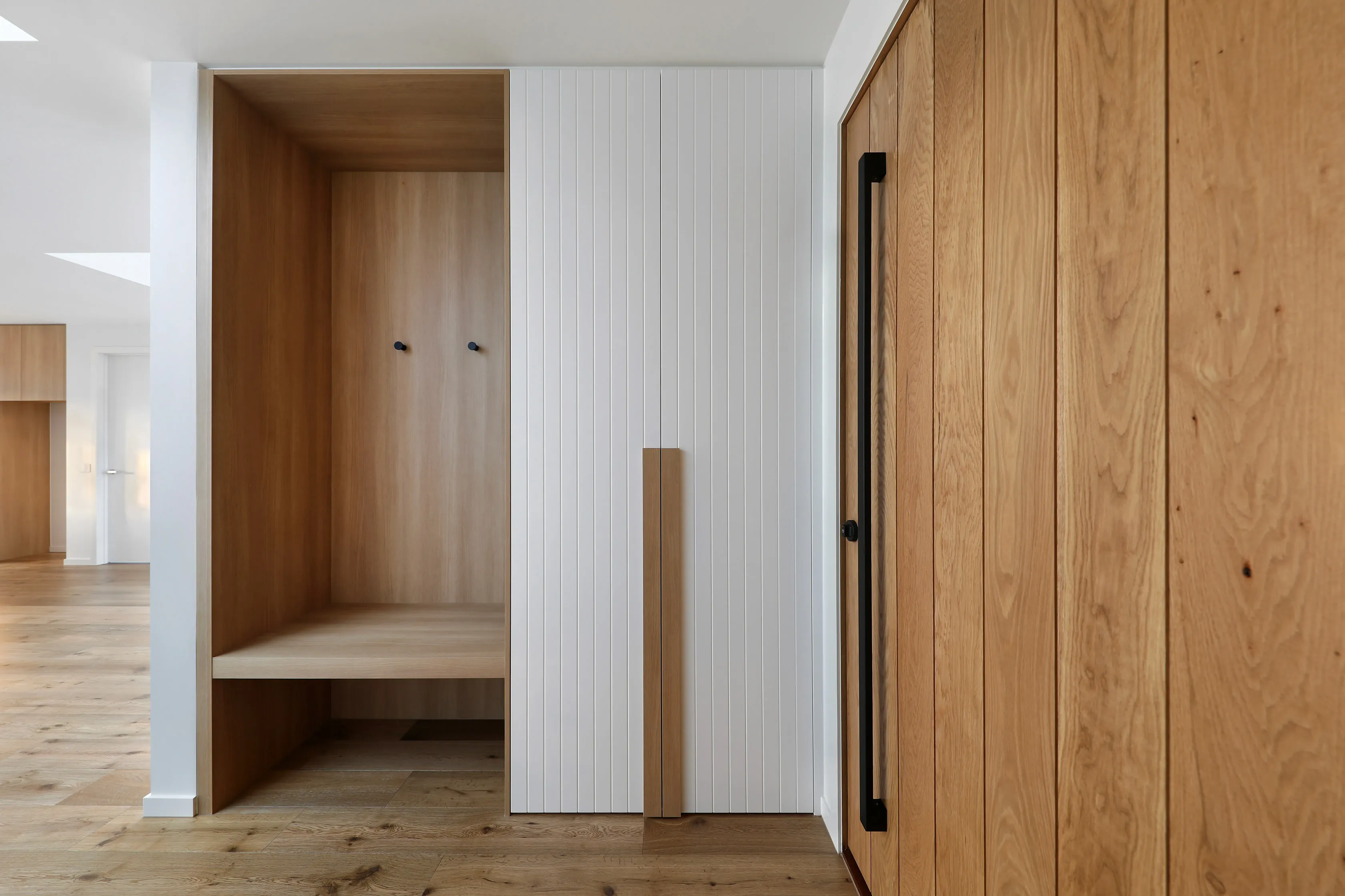 Entry mudroom with an oak-lined bench nook, white vertical-batten cabinetry, oak storage with a black handle and oak floors leading to the open living area