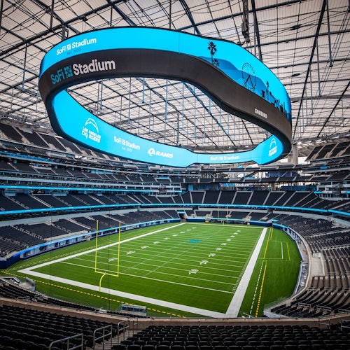 一个空荡荡的体育场，头顶是一个大型弯曲的电子屏幕，上面显示着“SoFi 体育场”，下面则是绿色的足球场。