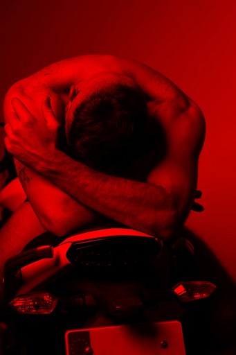 Hombre sentado en una motocicleta con los brazos cruzados, capturado en un estudio creativo con iluminación roja, mostrando una mezcla de moda y vanguardia a través de la fotografía profesional, resaltando la autenticidad y la sofisticación.