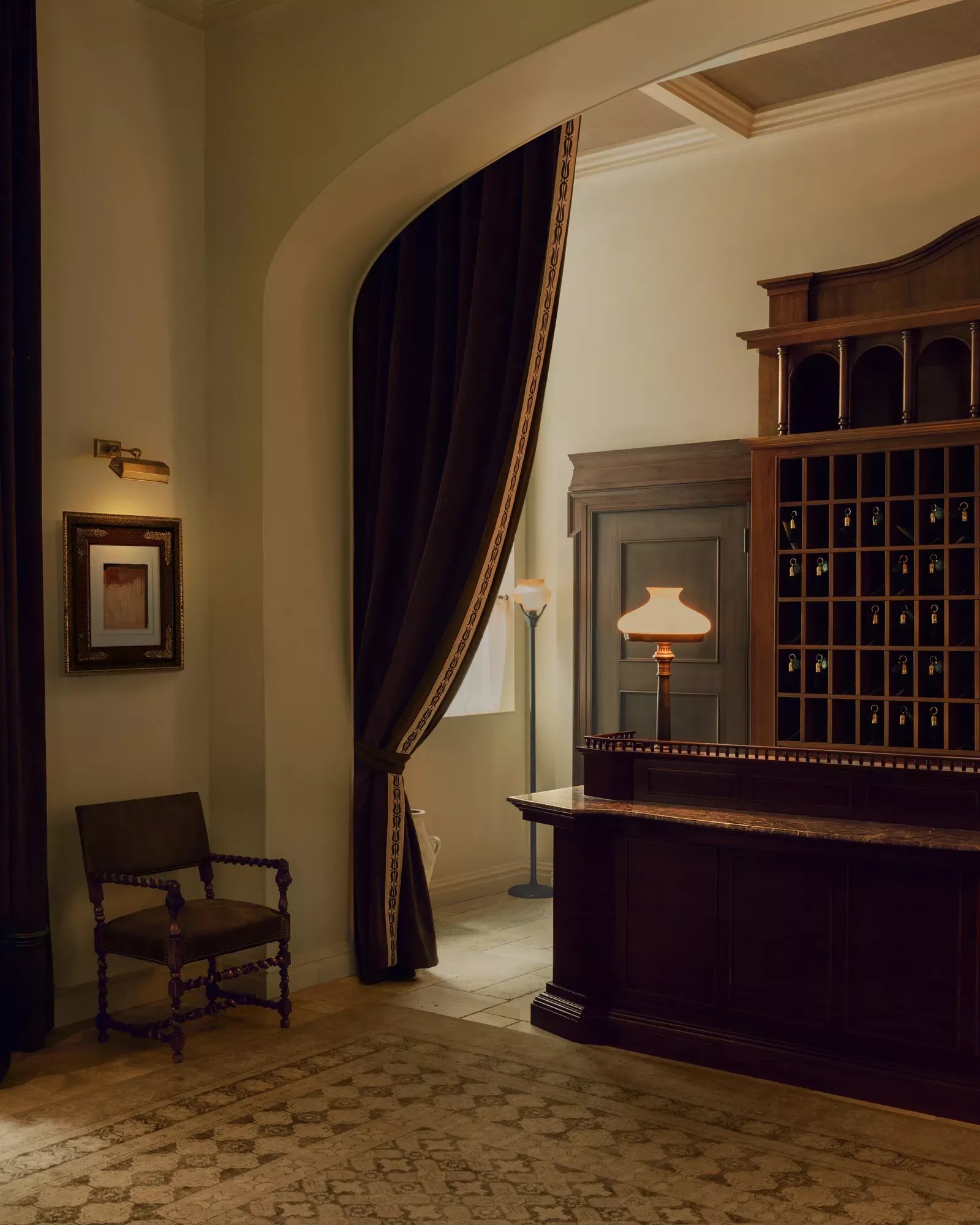 Elegant room framed by a large arch and curtains, with a lamp, chair, and dark wood counter.