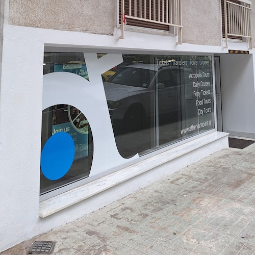 带有大型白色和蓝色标志的玻璃店面橱窗，上面展示了宣传各类旅游线路和服务的文字，并映出一辆汽车的倒影。