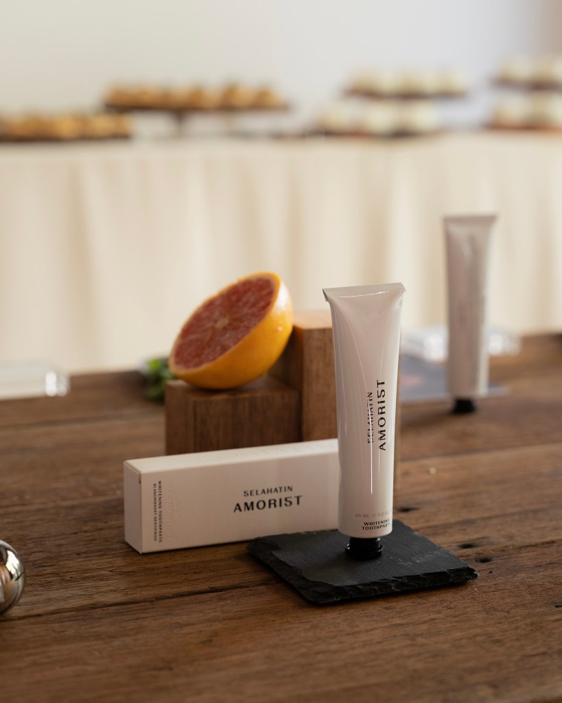 A tube of whitening toothpaste labeled 'AMORIST' by Selahatin, placed on a black slate coaster beside a halved grapefruit, with blurred dessert displays in the background.