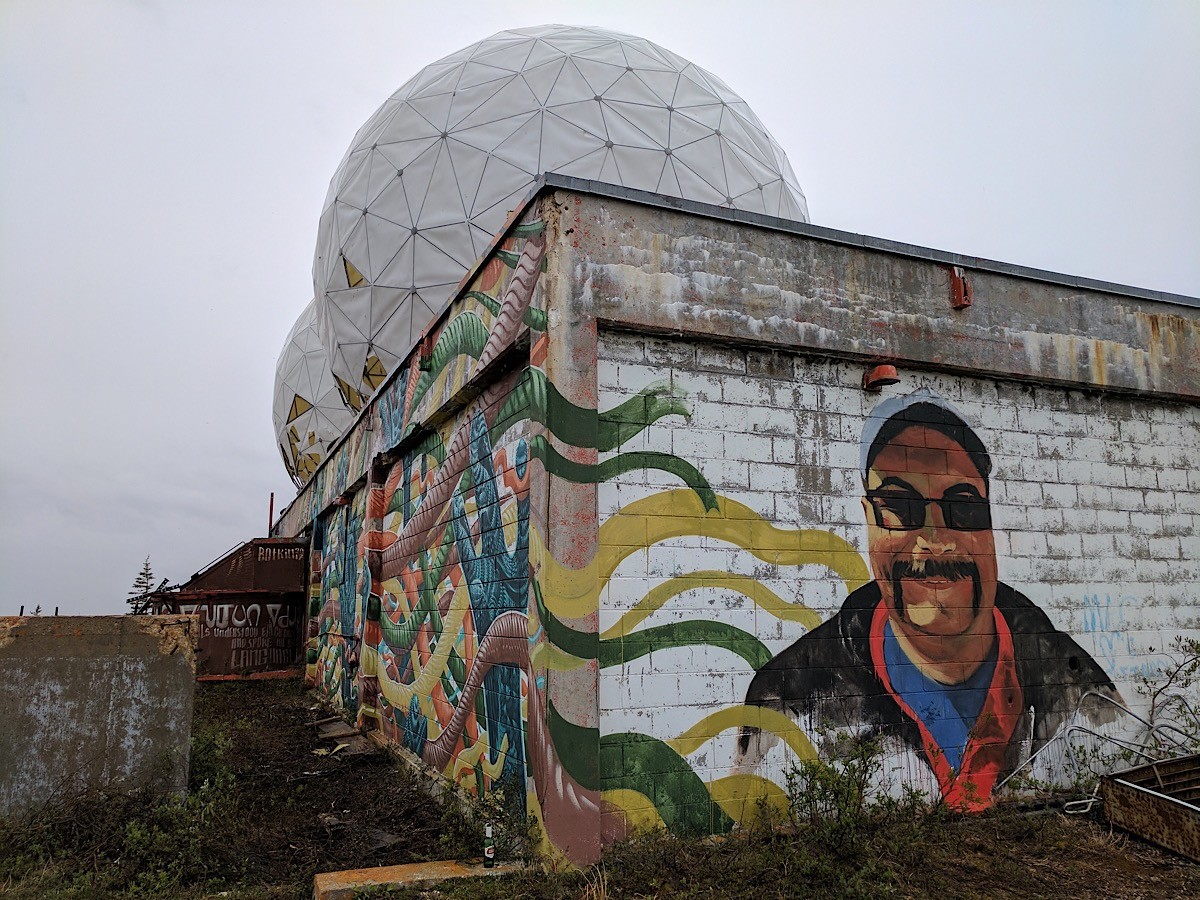 On a side wall beneath a radar dome, a smiling man in a cap and sunglasses, painted beside yellow and green bands.